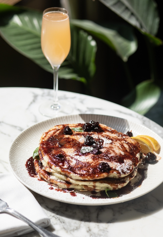 Stack of pancakes with berry sauce, powdered sugar, lemon wedge, and mimosa on marble table, leafy background.
