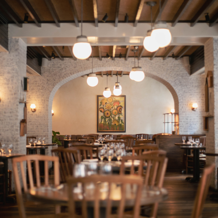 Cozy restaurant with wooden chairs, tables, stone walls, and arched ceilings. Warm lighting and floral artwork enhance the ambiance.