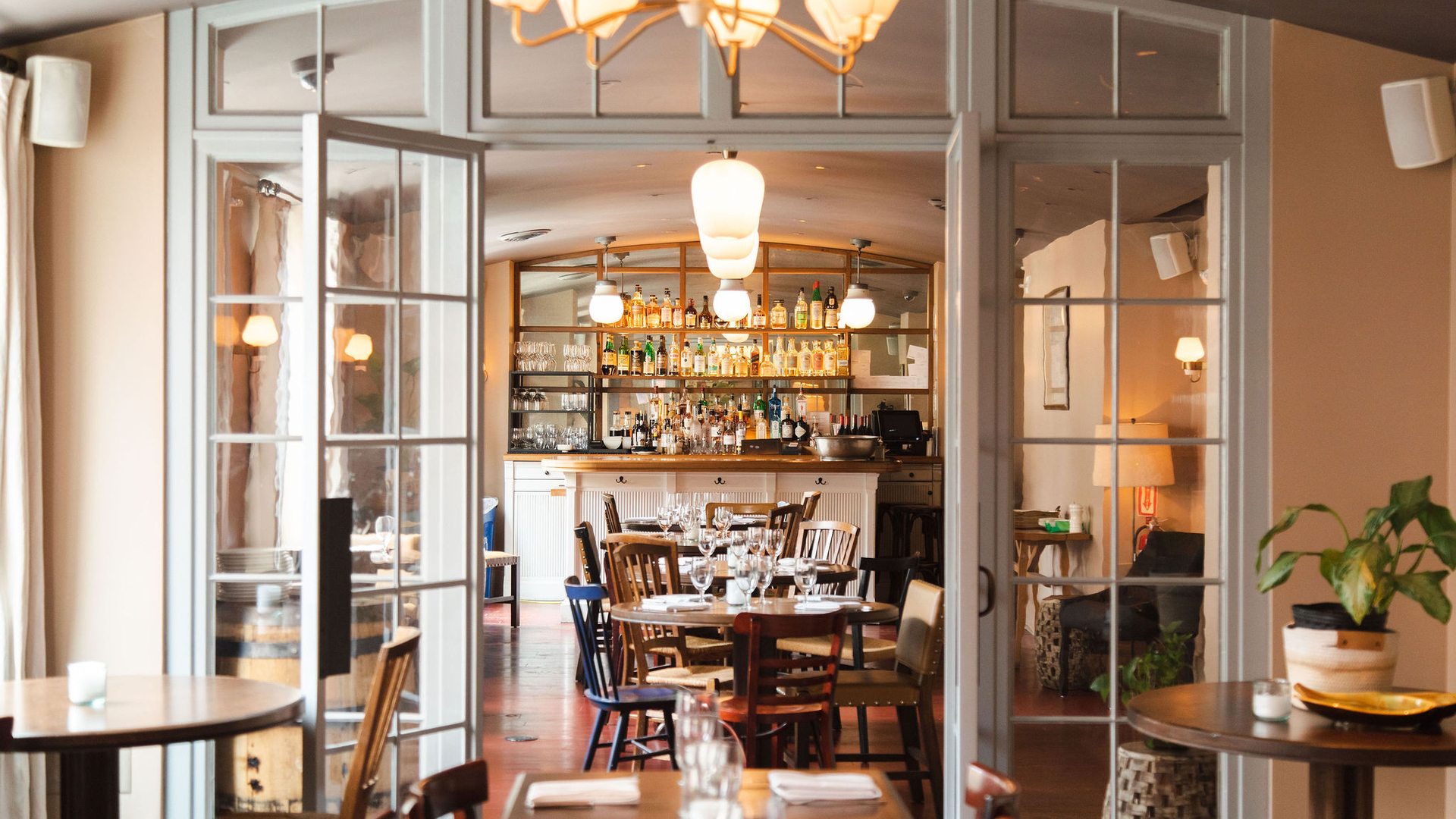 Cozy restaurant interior with glass doors, wooden tables, chairs, bar area, shelves, and plants.