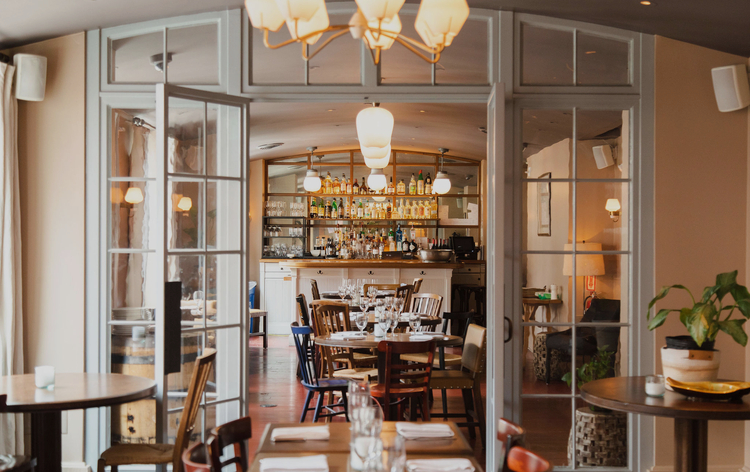 Modern restaurant with glass walls, open bar, wooden tables, and warm lighting.