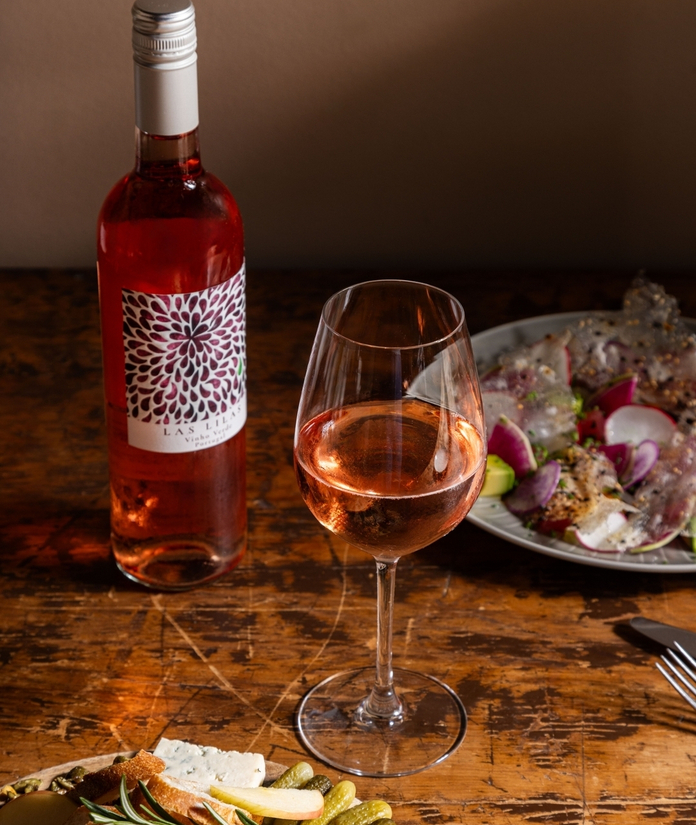 Pink wine and cheese platter on a rustic wooden table with a plate of food in the background.