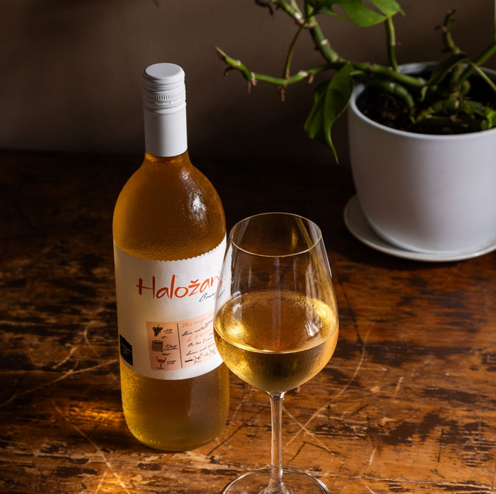 A bottle of Halozan white wine and a glass on a wooden table, with a potted plant in the background.
