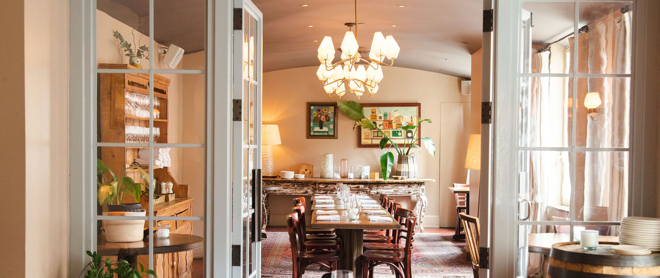 Warm, inviting dining room with large windows, chandelier, wooden tables, and barrel, open to a cozy kitchen.