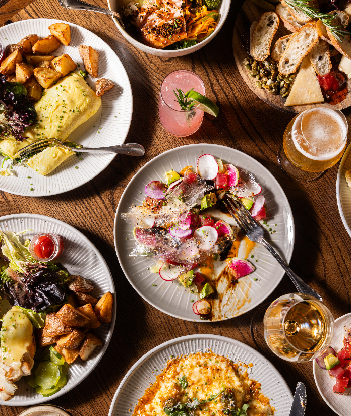 Overhead view of brunch dishes including eggs benedict, salads, charcuterie board, and cocktails on wooden table
