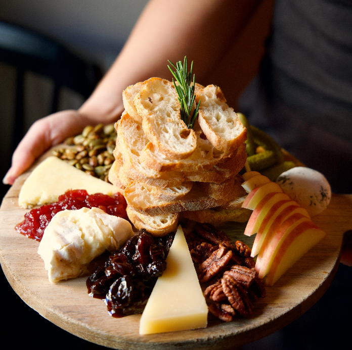 A round wooden cheese board being held up, arranged with sliced crusty bread, assorted cheeses, apple slices, dried fruit, pecans, pumpkin seeds, cranberry spread, and a sprig of rosemary.
