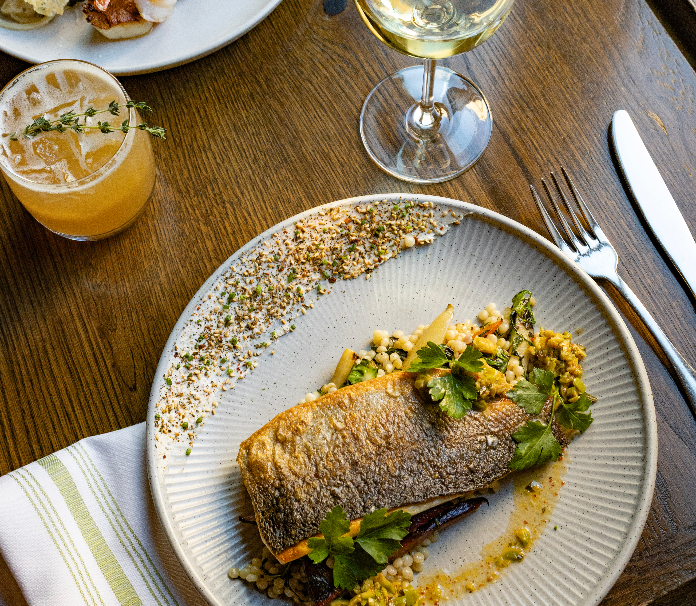 A plated fish entrée on a wooden table, featuring crispy-skinned fish over grains and vegetables, garnished with fresh herbs and sauce, with a glass of white wine and a cocktail nearby.