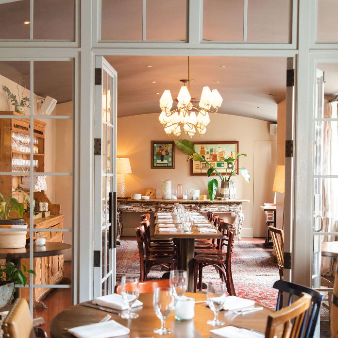 Warm, inviting dining room with large windows, chandelier, wooden tables, and barrel, open to a cozy kitchen.