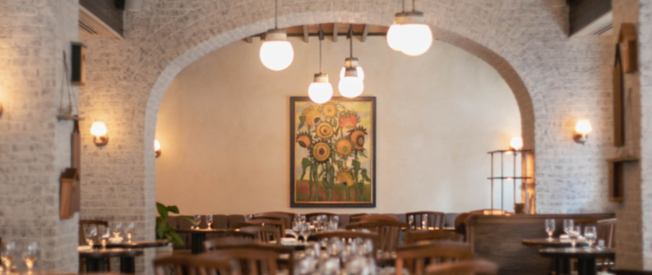 Cozy restaurant with wooden chairs, tables, stone walls, and arched ceilings. Warm lighting and floral artwork enhance the ambiance.